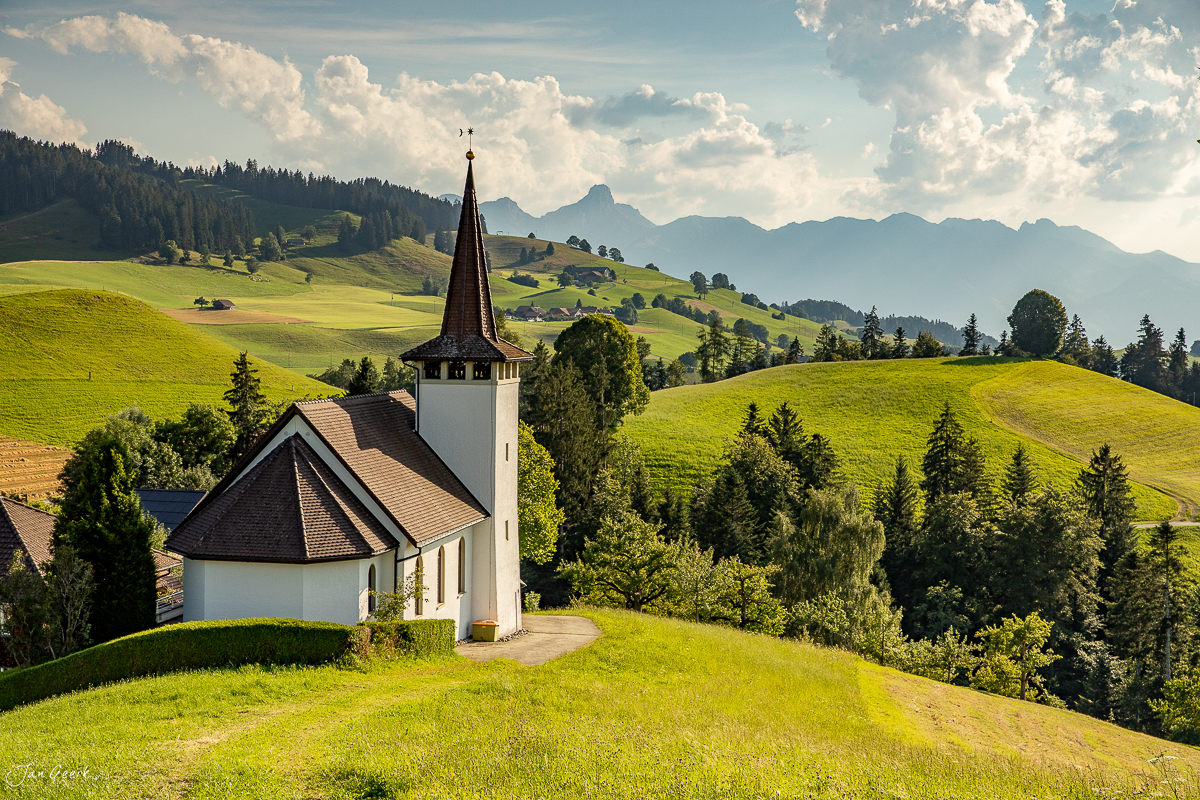 Kleine weiße Kirche mit Spitzturm in grüner Berglandschaft des Emmental mit Stockhorn im Hintergrund, Sommerlicht, Berner Oberland Schweiz – Jan Geerk Fotograf Basel