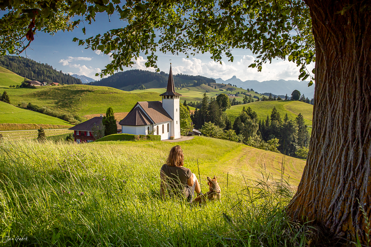 Person sitzt unter Buche und blickt auf Kirche im Emmental, grüne Sommerlandschaft mit Bergen, Schweiz – Jan Geerk Fotograf Basel