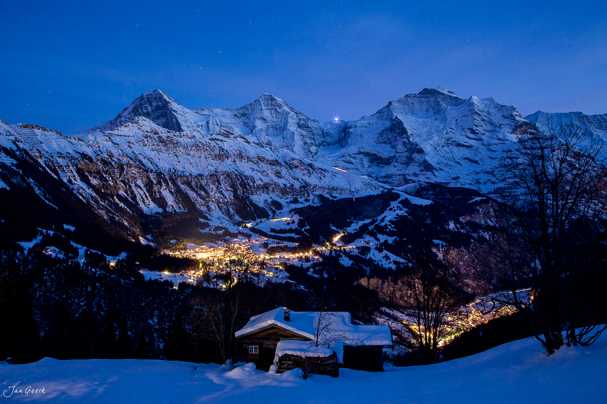 Verschneite Alphütte in Isenfluh mit beleuchteter Talstadt und schneebedeckten Berner Alpen bei Nacht – Schweiz Jan Geerk Fotograf Basel