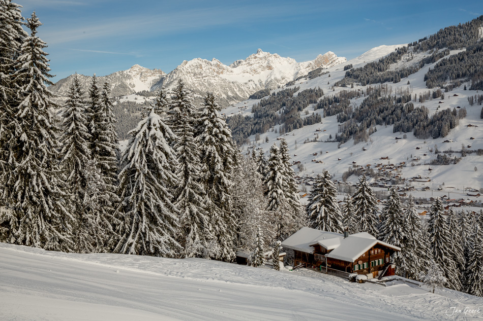 Verschneite Alphütte in der Lenk umgeben von schneebedeckten Lärchen, Berge im Hintergrund, Wintersonne, Berner Oberland Schweiz – Jan Geerk Fotograf Basel
