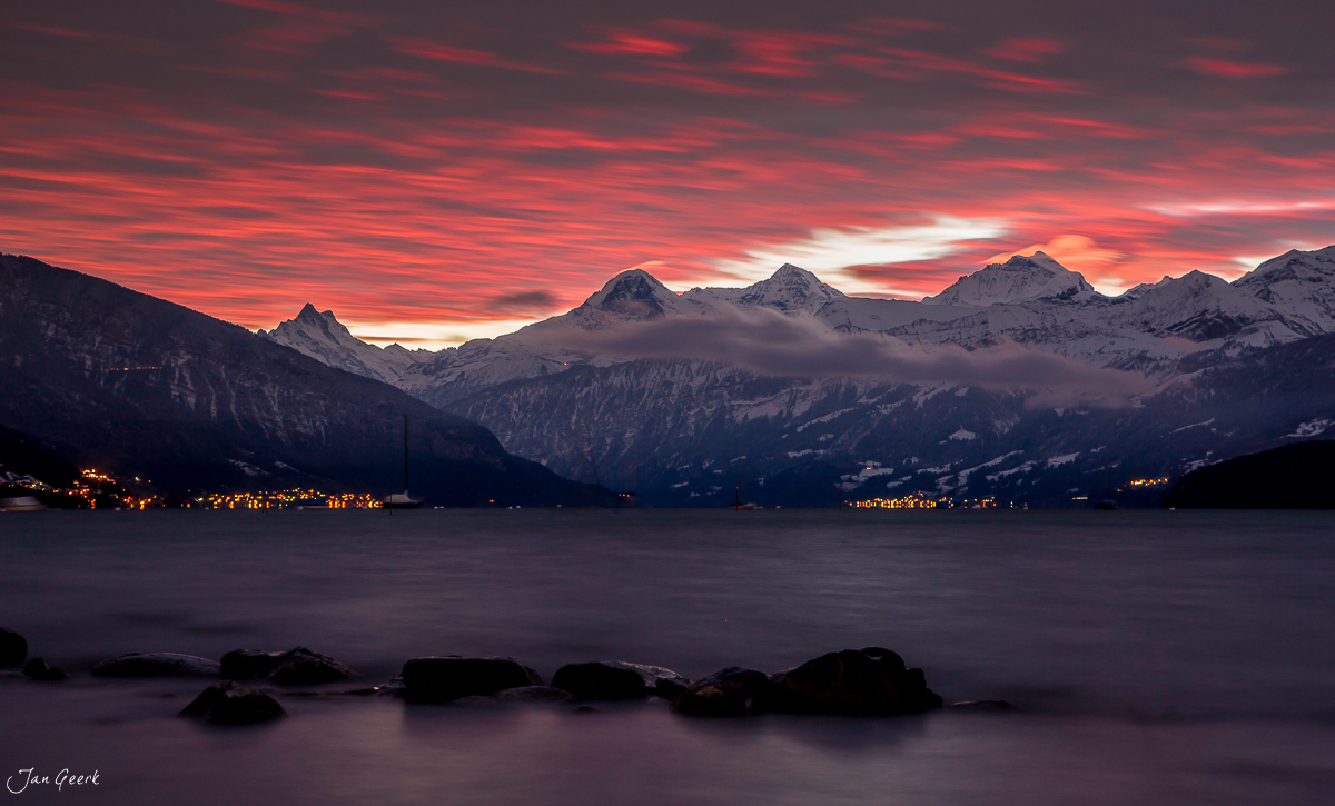 Dramatischer Sonnenuntergang über dem Thunersee mit schneebedeckten Alpen und beleuchteten Ufergemeinden im Winter – Schweiz, Jan Geerk Fotograf Basel