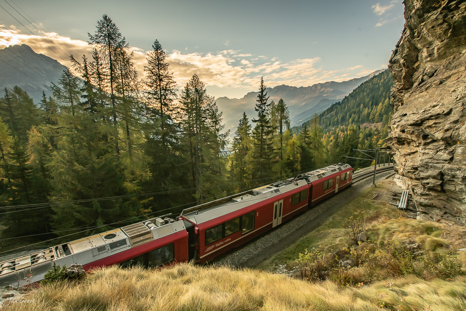 Rote Zahnradbahn der Rhätischen Bahn in Poschiavo mit Herbstlandschaft, Lärchenwälder und Felswand bei Kantlicht – Schweiz von Jan Geerk Fotograf Basel