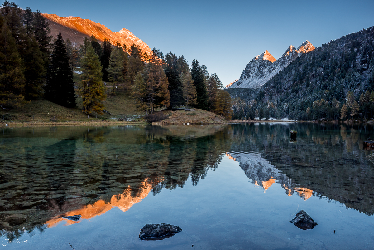 Bergsee mit Spiegelung herbstlicher Lärchen und beleuchteter Berggipfel im Engadin Graubünden Schweiz – Jan Geerk Fotograf Basel