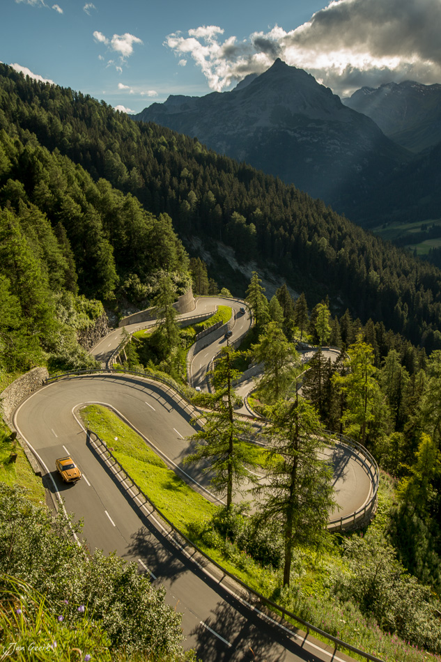 Serpentinen-Bergstrasse durch Lärchenwälder im Engadin, Sommeraufnahme mit gelben Taxifahrzeug, Schweiz – Jan Geerk Fotograf Basel
