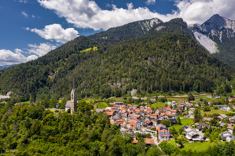 Luftaufnahme des Dorfes Tamins im Churer Rheintal mit Kirche, grünen Wiesen und bewaldeten Bergen unter blauem Himmel – Jan Geerk Fotograf Basel