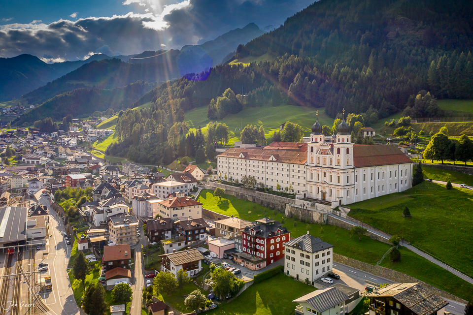 Luftaufnahme des Benediktinerklosters Disentis mit Alpenpanorama, Sonnenstrahlen und grünen Wiesen, Graubünden Schweiz – Jan Geerk Fotograf Basel