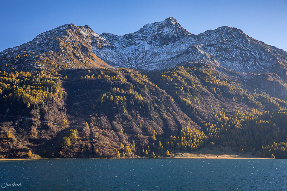 Bergsee mit Lärchenwäldern im Herbst, schneebedeckte Berggipfel, goldenes Licht in Graubünden Schweiz – Jan Geerk Fotograf Basel