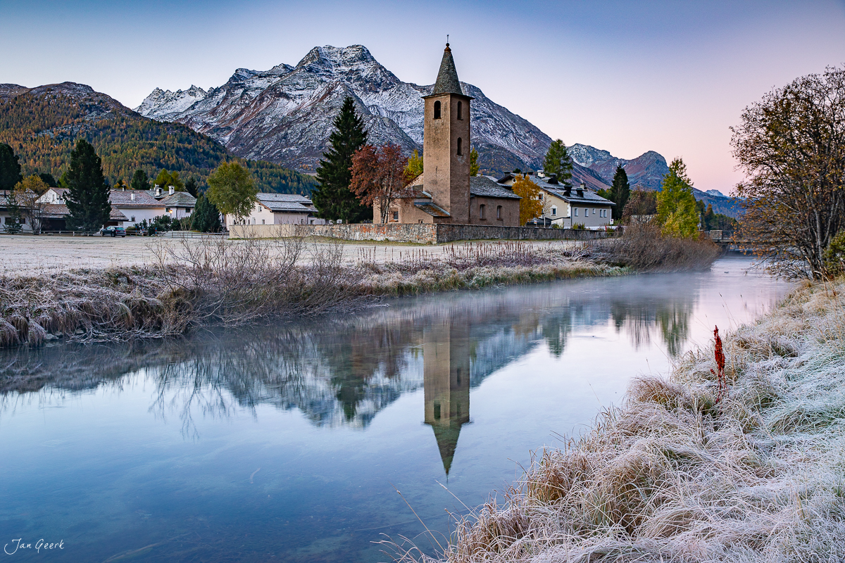 Kirche mit Kirchturm spiegelt sich im ruhigen Fluss, schneebedeckte Berge im Hintergrund, Herbststimmung in der Schweiz – Jan Geerk Fotograf Basel