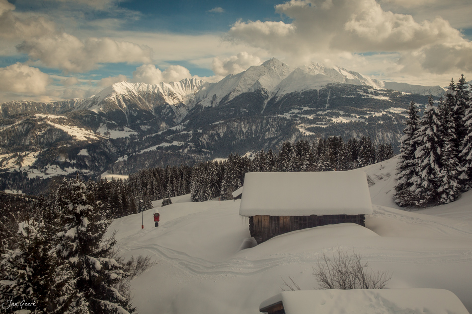 Schneebedeckte Alphütte in Laax mit verschneiten Bergen und Fichten im Winterlicht, Graubünden Schweiz – Jan Geerk Fotograf Basel