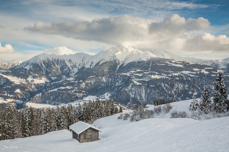 Schneebedeckte Almhütte in Laax mit verschneiten Bergen und Nadelwald im Hintergrund, Winterstimmung in Graubünden Schweiz – Jan Geerk Fotograf Basel