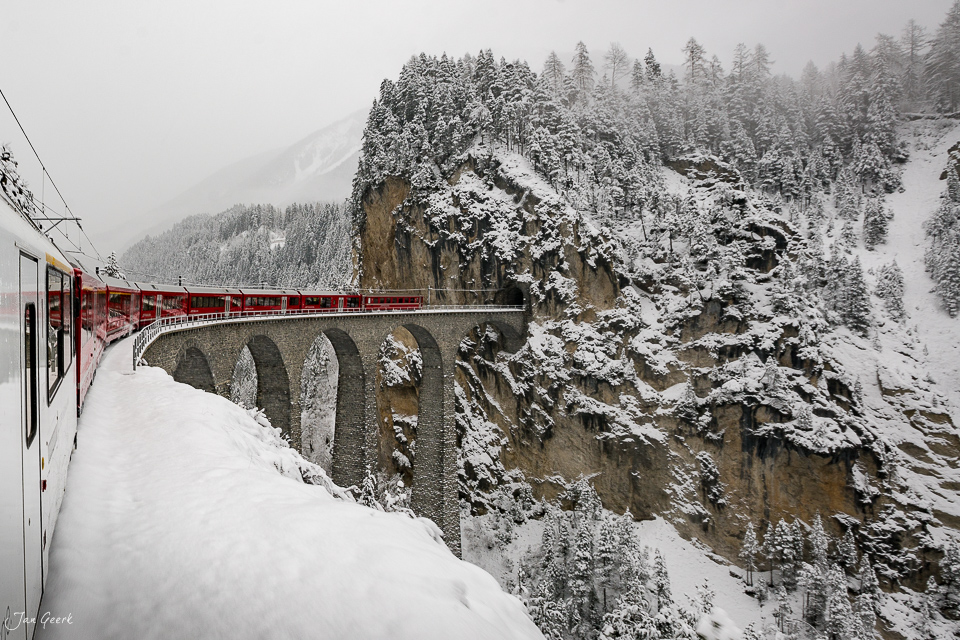 Landwasserviadukt im Winter mit rotem Zug, schneebedeckte Felsen und Wald, Graubünden Schweiz – Jan Geerk Fotograf Basel