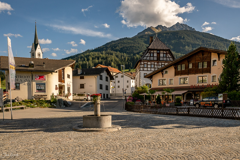 Dorfplatz mit Kirche und traditionellen Häusern in Savognin, Berge im Hintergrund, Sommer, Graubünden Schweiz – Jan Geerk Fotograf Basel