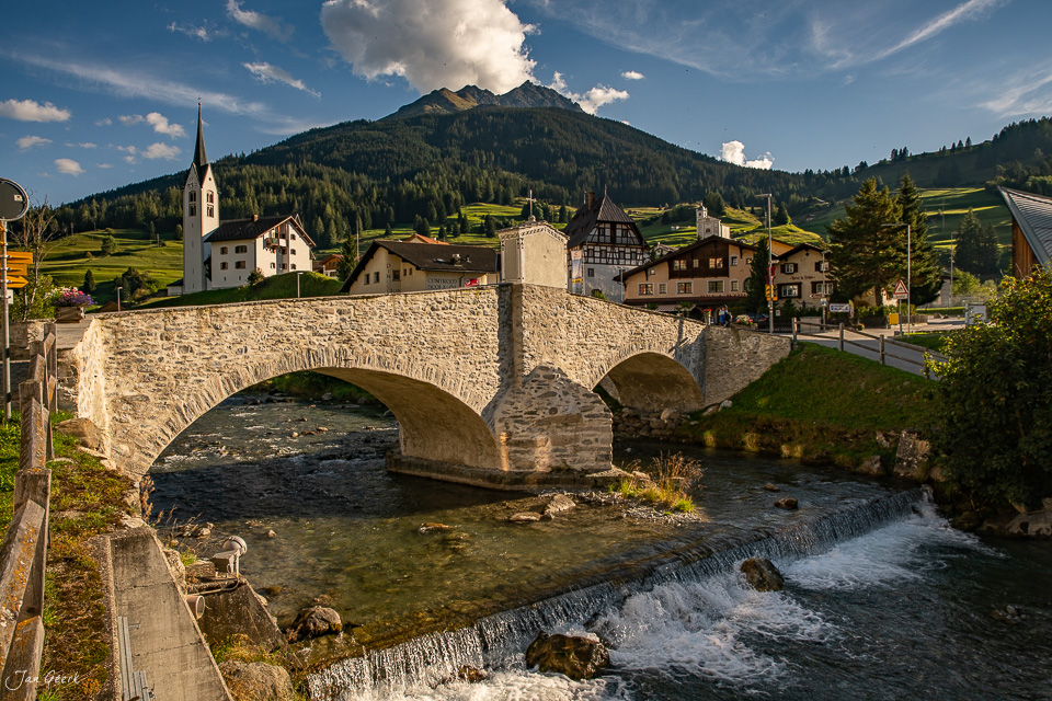 Steinerne Brücke über Fluss mit Bergdorf Savognin im Hintergrund, Sommerliche Alpenlandschaft Graubündens Schweiz – Jan Geerk Fotograf Basel