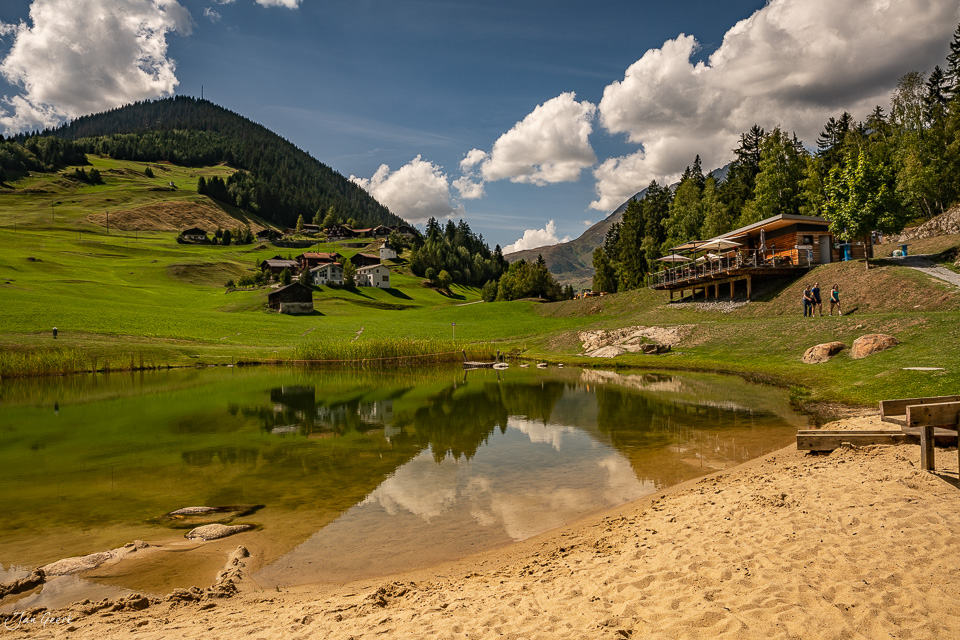 Alpensee mit Berghütten und grünen Wiesen in Sedrun unter blauem Himmel mit Sonnenschein, Graubünden Schweiz – Jan Geerk Fotograf Basel