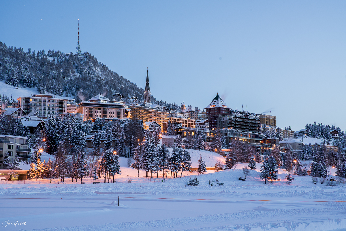 Winterliches Dorf St. Moritz mit schneebedeckten Häusern und beleuchteten Hotels am gefrorenen See in der Abenddämmerung, Graubünden Schweiz – Jan Geerk Fotograf Basel