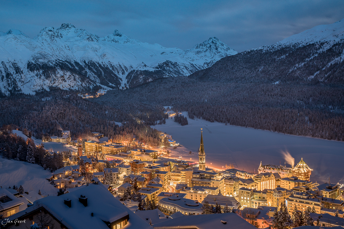 Schneebedecktes Dorf St. Moritz mit beleuchteten Gebäuden, verschneiten Bergen und gefrorenem See in der Winterdämmerung – Graubünden, Schweiz – Jan Geerk Fotograf Basel