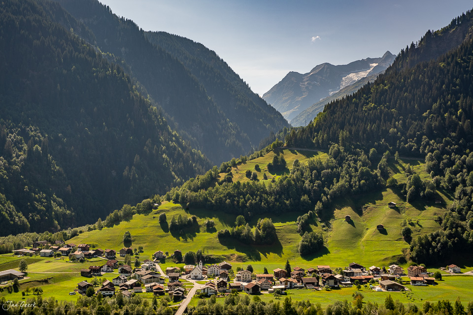 Alpental mit Bergdorf, grünen Wiesen und Nadelwäldern unter blauem Himmel, Schweiz – Jan Geerk Fotograf Basel