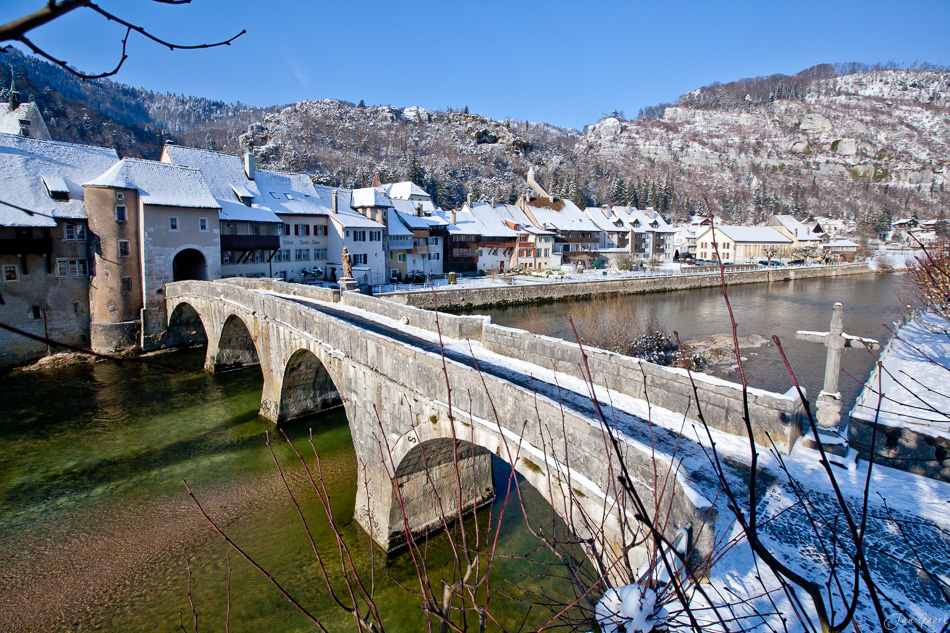 Steinbrücke über Fluss schneebedeckte Häuser Winter Franches Montagnes Jura Schweiz Jan Geerk Fotograf Basel