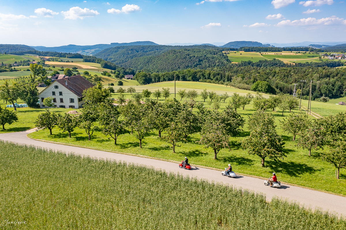 Kinder auf Vespas fahren eine Landstrasse in Häfelfingen entlang, umgeben von grünen Wiesen und Obstbäumen, Baselland Schweiz – Jan Geerk Fotograf Basel
