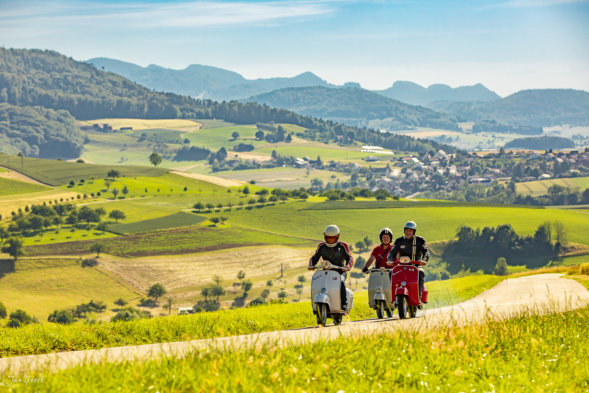 Drei Vespa-Fahrer auf Landstraße im Baselbiet mit grünen Hügeln und Bergpanorama, Sommerlicht, Schweiz – Jan Geerk Fotograf Basel