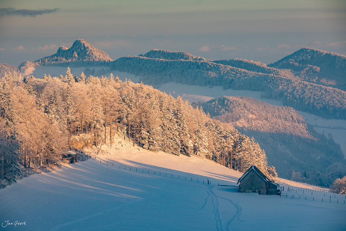 Verschneite Waldweid mit traditionellem Stall, schneebedeckten Fichten und Bergen im Morgenlicht, Baselland Winter Schweiz – Jan Geerk Fotograf Basel