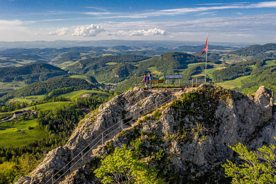 Aussichtsplattform mit Schweizer Flagge auf dem Belchen, grüne Hügellandschaft im Frühling, Luftaufnahme Baselland Schweiz – Jan Geerk Fotograf Basel