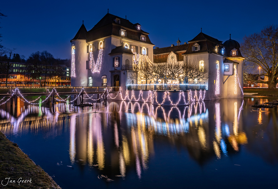 Schloss Bottmingen mit festlicher Weihnachtsbeleuchtung in der Abenddämmerung, gespiegelt im Wasser, Baselland Schweiz – Jan Geerk Fotograf Basel