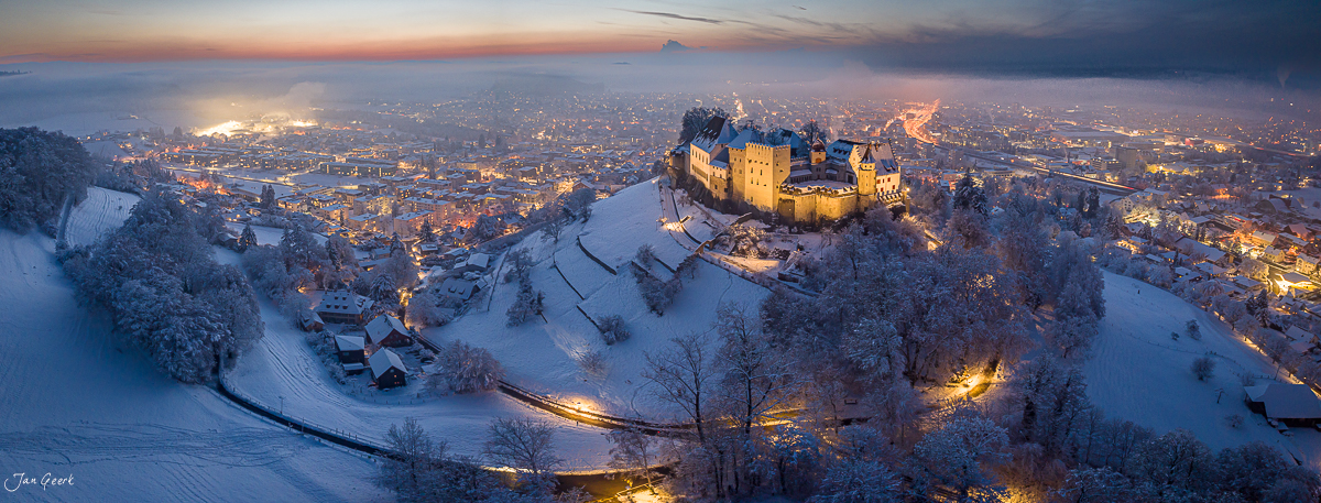 Schloss Lenzburg winterliche Luftaufnahme bei Dämmerung mit verschneiter Landschaft und beleuchteter Stadt im Hintergrund Schweiz Jan Geerk Fotograf Basel