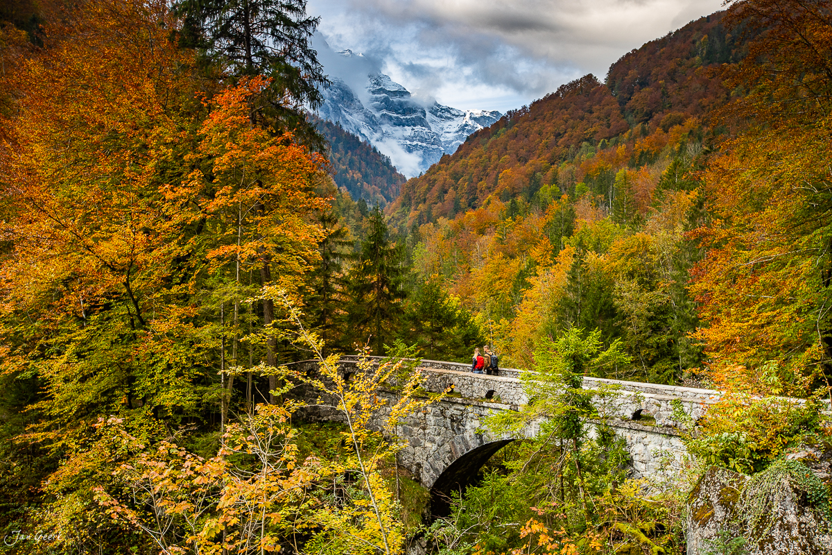 Herbstliche Berglandschaft am Klöntalersee mit schneebedecktem Alpengipfel, Steinbrücke und goldenem Laubwald in Glarus, Schweiz – Jan Geerk Fotograf Basel