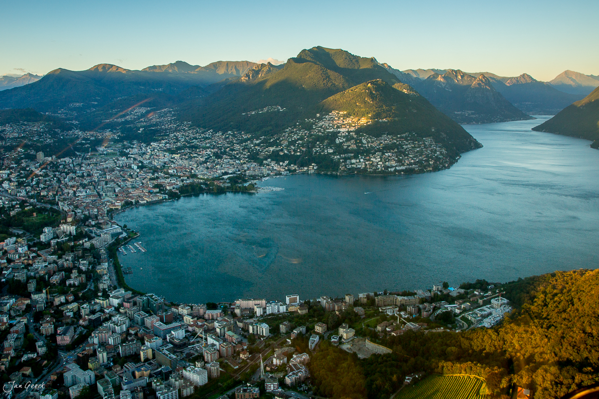 Luftaufnahme Lugano mit türkisem See, umliegenden Bergen und beleuchteter Stadt in goldenem Abendlicht Tessin Schweiz – Jan Geerk Fotograf Basel
