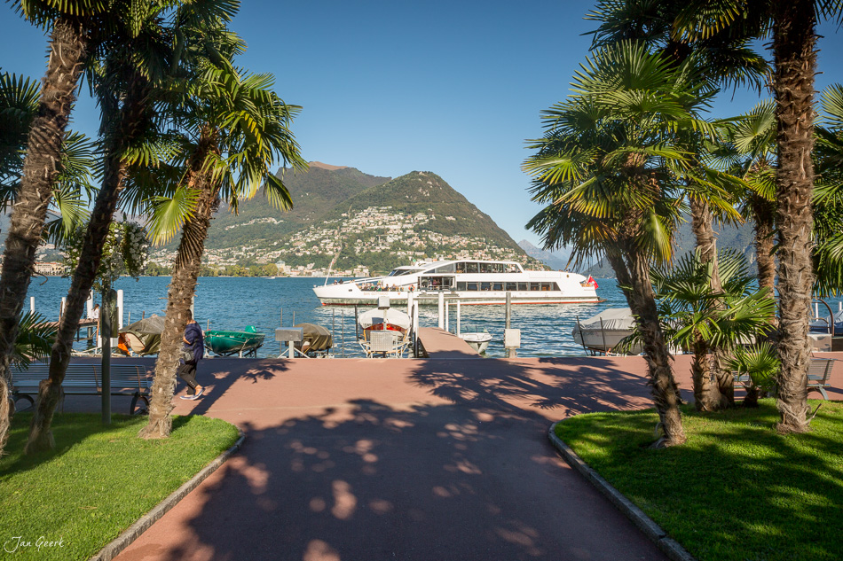 Seeufer mit Palmen, Fährschiff und Berglandschaft in Lugano, sonniger Sommertag, Tessin Schweiz – Jan Geerk Fotograf Basel