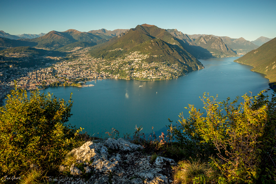 Blick auf Lugano mit türkisblauem See, umgeben von grünen Bergen und bebauten Hängen an einem sonnigen Tag in der Schweiz – Jan Geerk Fotograf Basel