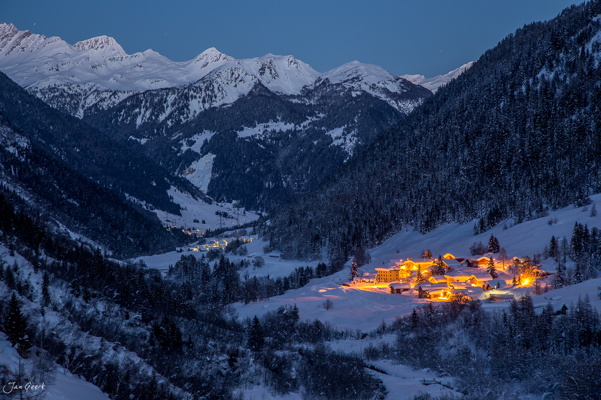 Verschneite Berghäuser in Bedretto mit beleuchteten Chalets und schneebedeckten Gipfeln der Südalpen im Tessin Schweiz – Jan Geerk Fotograf Basel