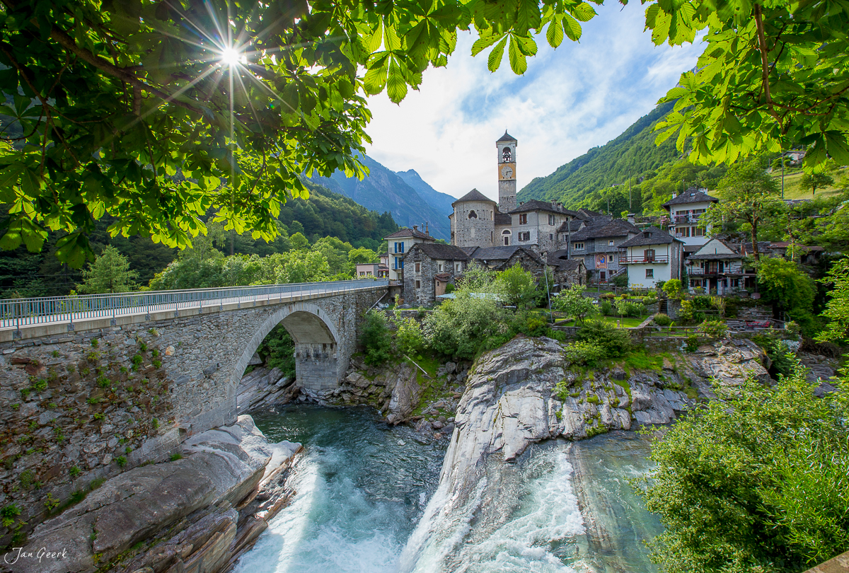 Steinbrücke über türkises Wasser im Bleniotal mit Kirchturm und Bergdorf unter Frühjahrssonnenschaft, Tessin Schweiz – Jan Geerk Fotograf Basel