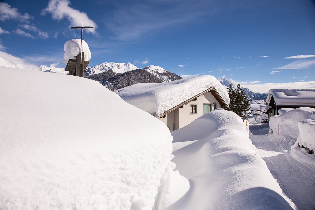Verschneite Bergkapelle mit Kreuz in Bedretto, verschneite Alpenhäuser, Wintersonne, Tessin Schweiz – Jan Geerk Fotograf Basel