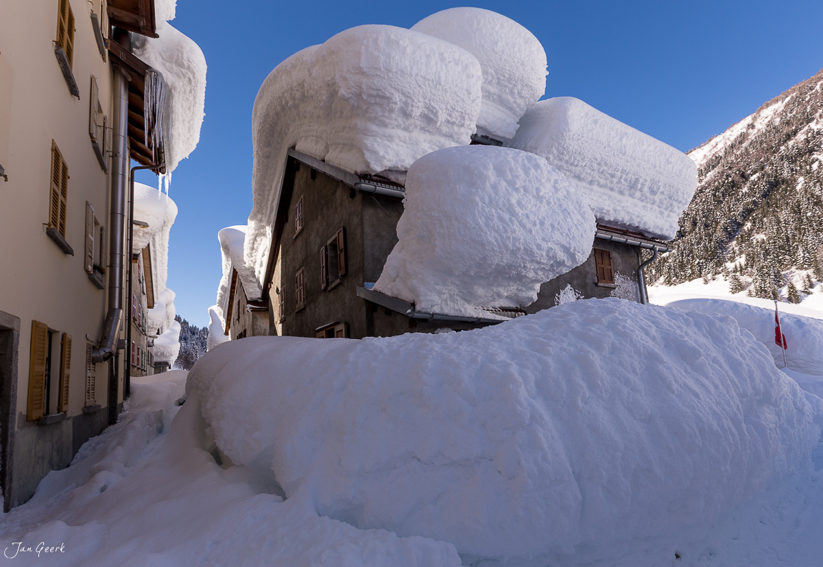 Schneebedeckte Häuser und Dorfstrasse in Airolo, verschneite Winterszene in der Schweiz mit mächtigen Schneemassen auf den Dächern, sonnig und klar, Tessin – Jan Geerk Fotograf Basel