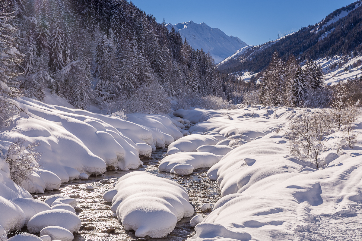 Schneebedeckter Fluss mit verschneiten Steinen im Bedretto Winterlandschaft Tessin Schweiz – Jan Geerk Fotograf Basel