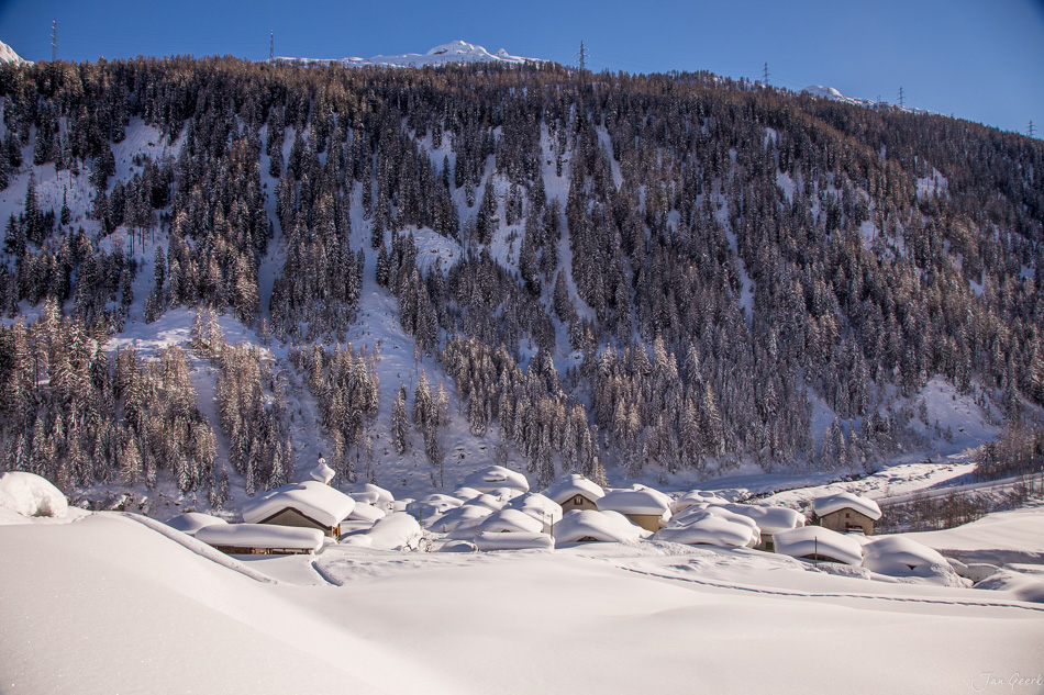Schneebedeckte Berghütten in Bedretto mit verschneitem Lärchenwald und Alpengipfel im Hintergrund, Tessin Schweiz – Jan Geerk Fotograf Basel