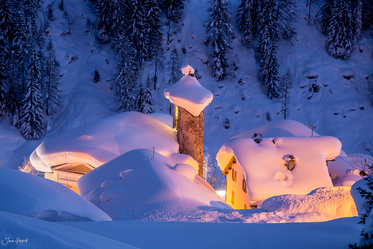 Verschneite Villa mit beleuchteter Kirche in Bedretto, Winterabendstimmung, Nordtessin Schweiz – Jan Geerk Fotograf Basel