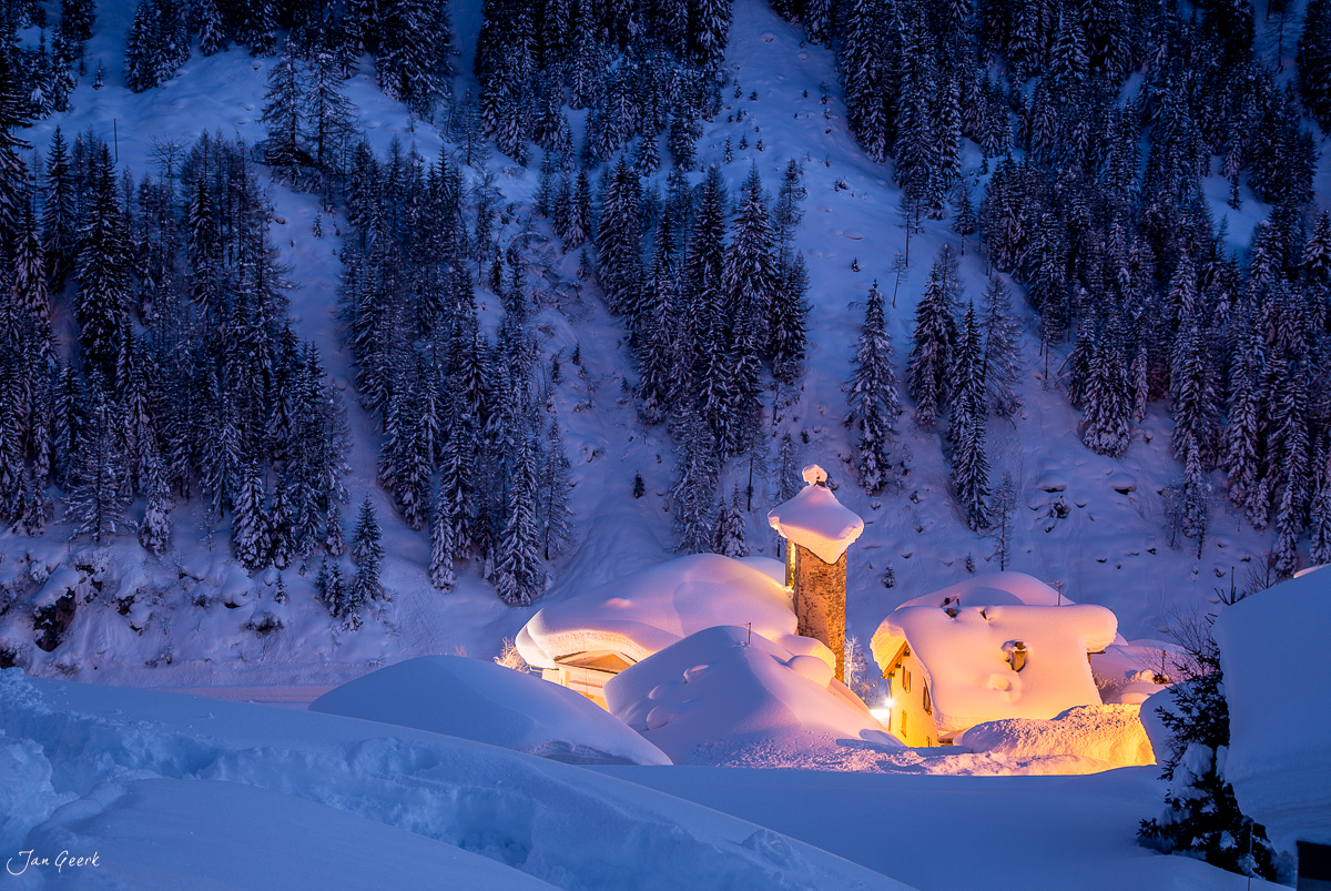 Verschneite Villa mit leuchtender Beleuchtung in winterlicher Berglandschaft umgeben von schneebedeckten Fichten im Tessin Schweiz Jan Geerk Fotograf Basel