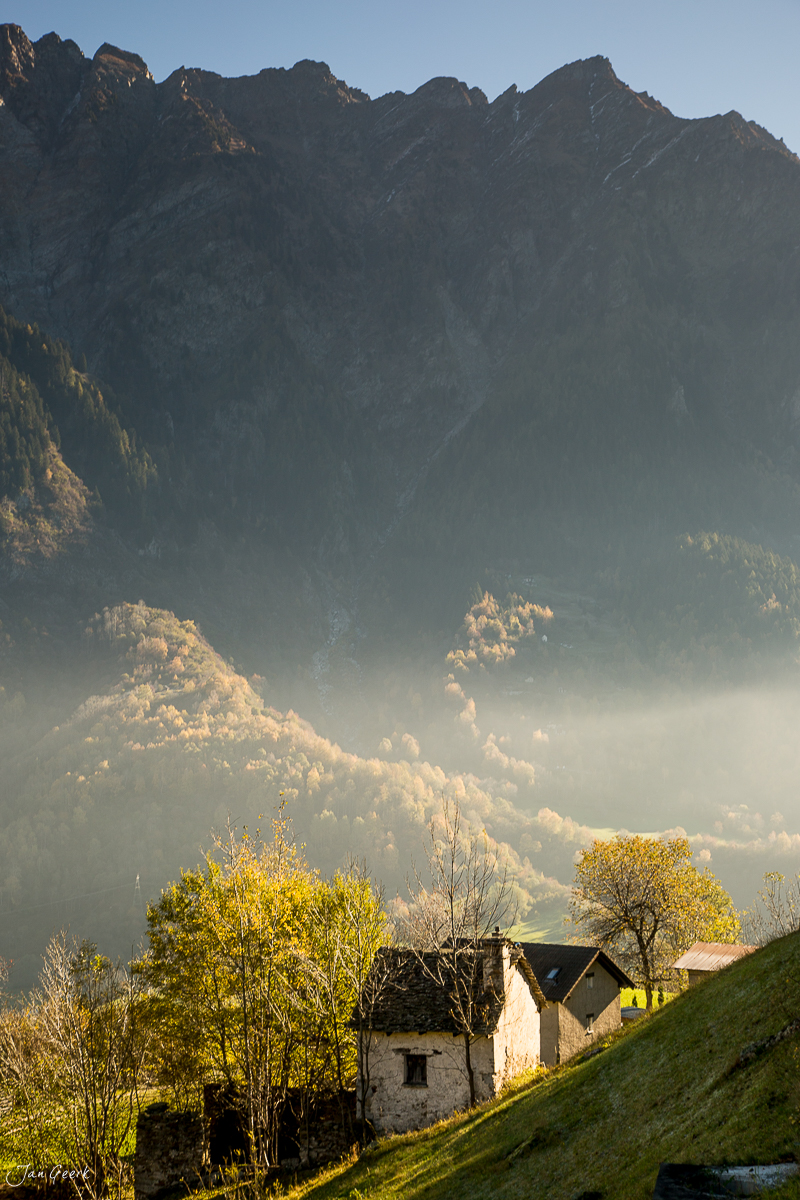 Alpines Tal mit traditionellem Bauernhaus, Herbstbäumen und nebligen Bergen im Tessin Schweiz – Jan Geerk Fotograf Basel