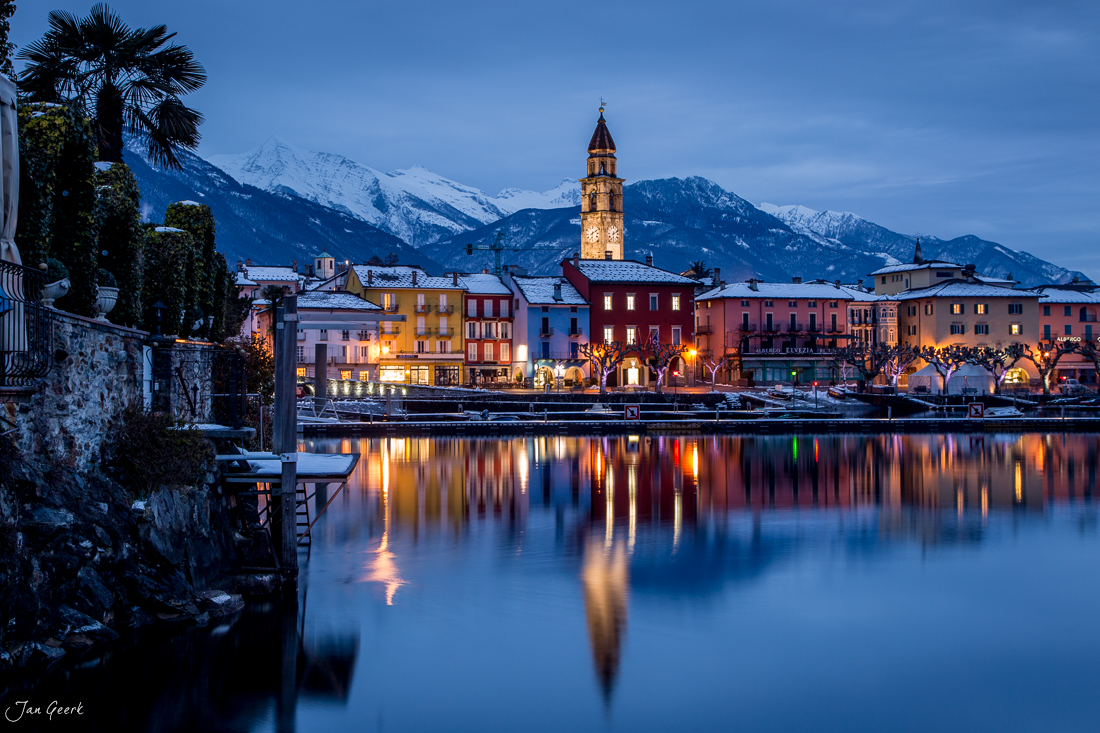Ascona Ufer mit schneebedeckten Bergen in Winternacht, beleuchtete Häuser spiegeln sich im Lago Maggiore, Tessin Schweiz – Jan Geerk Fotograf Basel