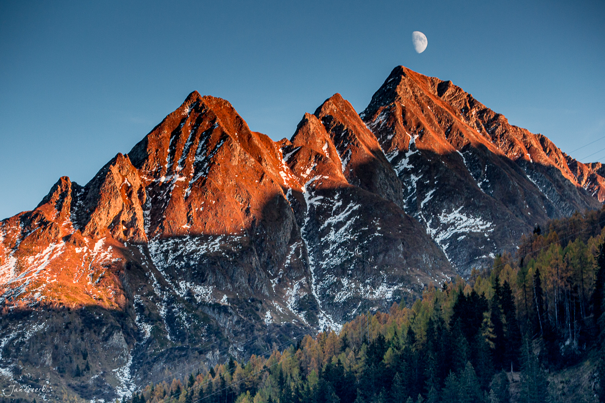 Schneebedeckte Berggipfel im Tessin mit Herbstlärchen und Mond am blauen Himmel, Schweiz – Jan Geerk Fotograf Basel