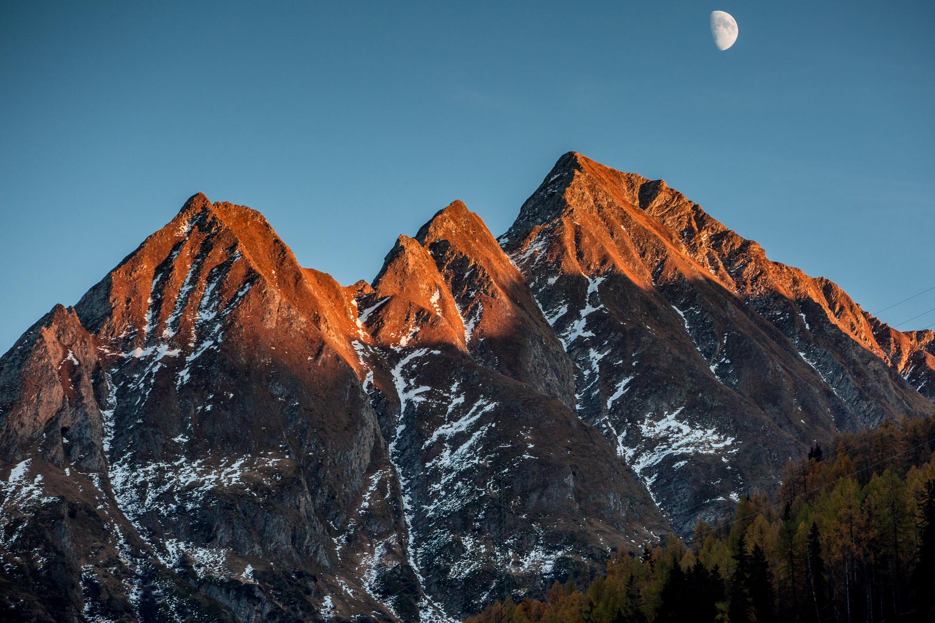 Verschneite Berggipfel im Tessin mit goldener Herbstbeleuchtung und Mond am Tag, Schweiz – Jan Geerk Fotograf Basel