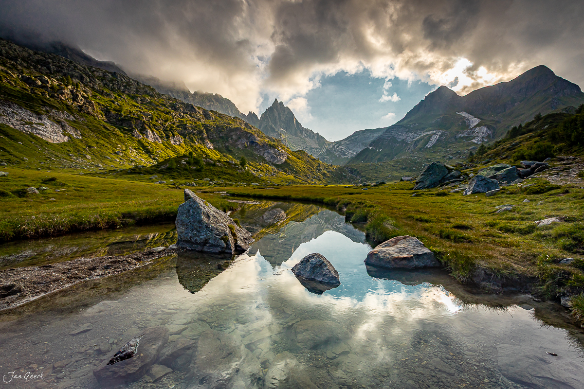 Alpiner See mit Spiegelung, umgeben von grünen Wiesen und dramatischen Bergen im Tessin, Sommer – Jan Geerk Fotograf Basel