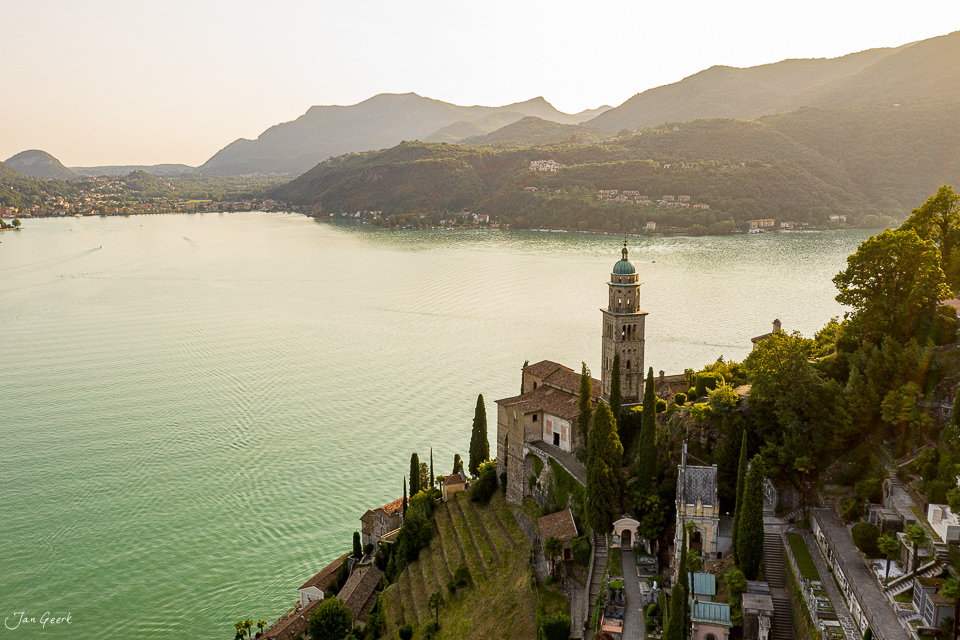 Kirchturm und Friedhof an steiler Seeufer-Klippe oberhalb des Sees mit bewaldeten Bergen im Morgenlicht, Tessin Schweiz – Jan Geerk Fotograf Basel