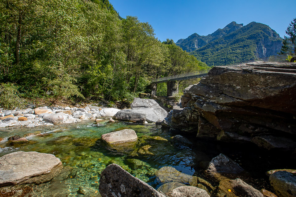 Kristallklarer Gebirgsbach Verzascatal mit Steinbrücke, grünen Laubbäumen und Bergen im Tessin Sommer Schweiz – Jan Geerk Fotograf Basel