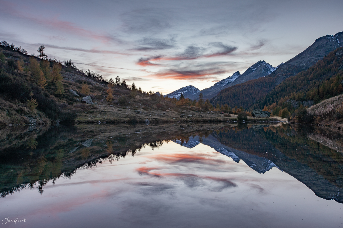Spiegelglatter See im Lötschental mit schneebedeckten Bergen, Lärchenwäldern und dramatischem Sonnenuntergangshimmel in Rot und Orange, Wallis Schweiz – Jan Geerk Fotograf Basel