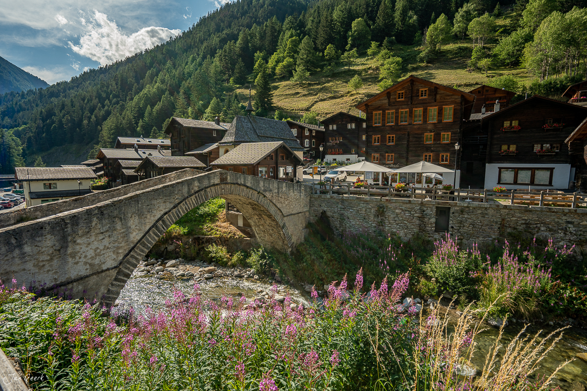 Steinbrücke über Bach mit Alpendorf, Lärchenwäldern und lilafarbenen Blüten im Sommer Wallis Schweiz – Jan Geerk Fotograf Basel