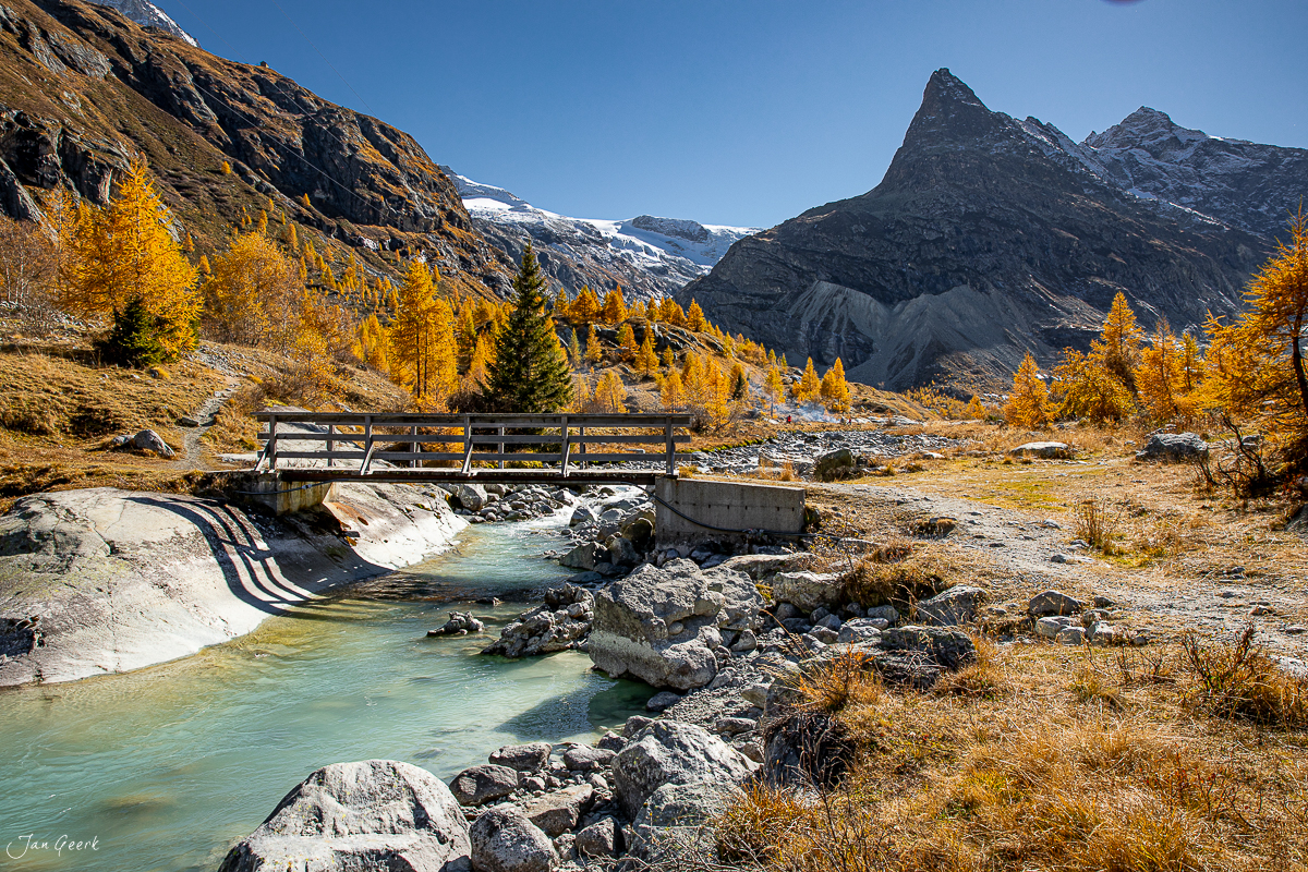 Holzbrücke über türkisblauer Bergbach mit golden gefärbten Lärchen im Herbst bei Ferpecle Wallis Schweiz – Jan Geerk Fotograf Basel